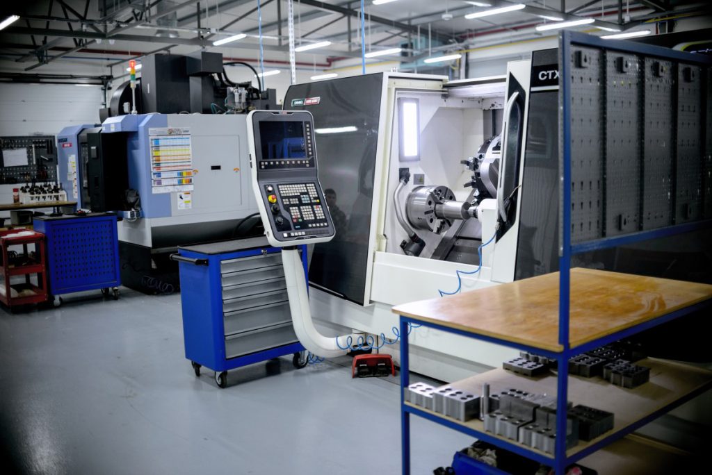 A production hall with a CNC lathe, control panel, blue tool trolley and workbench with components.