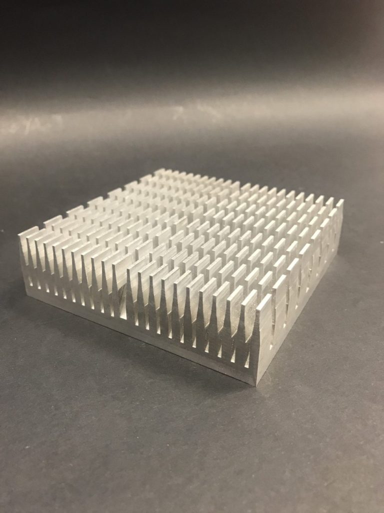 A silver square aluminium heat sink with dense vertical fins, positioned at an angle against a dark grey background.