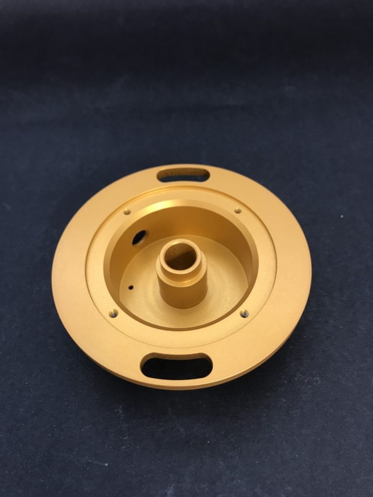 A round gold-coloured metal component milled on a CNC machine, with mounting holes and a sleeve, lying against a dark background.