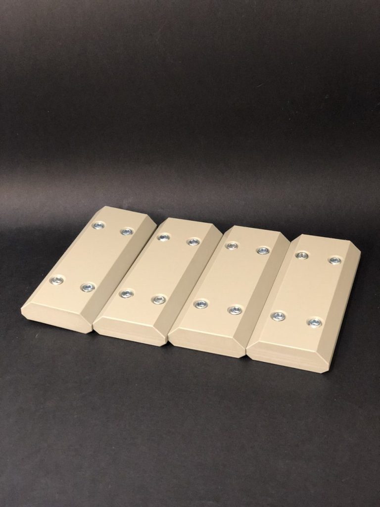 Four beige milled components with mounting holes and chamfered edges lying side by side against a black background.