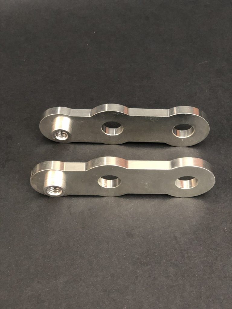 Two milled metal components with two holes and a threaded sleeve, lying on a matt black background.