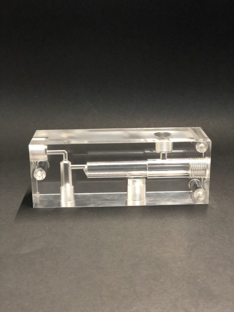 A transparent acrylic block with intricate channels milled inside, standing against a dark background.
