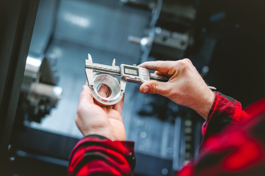 A worker's hands measuring a turned metal ring with digital callipers inside a CNC machine enclosure.