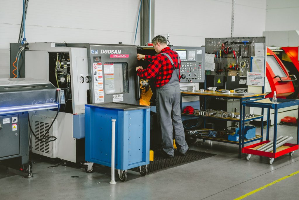 A worker in a checked shirt operates a CNC lathe in a bright production hall with tool trolleys.