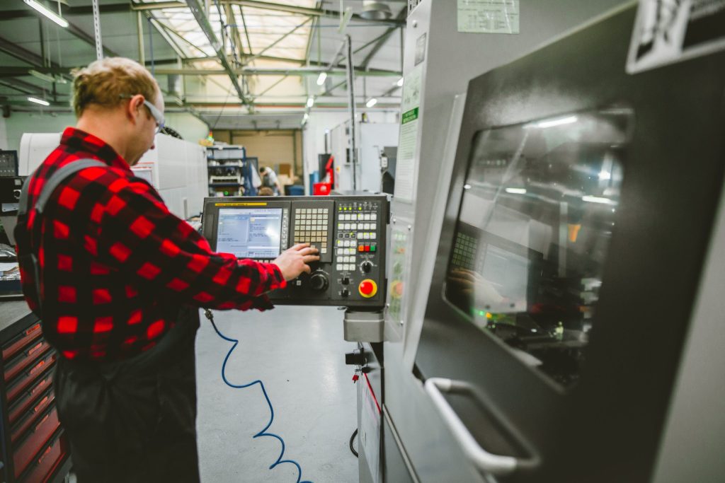 An operator in a red-and-black checked shirt operates the control panel of a CNC machine in a production hall.