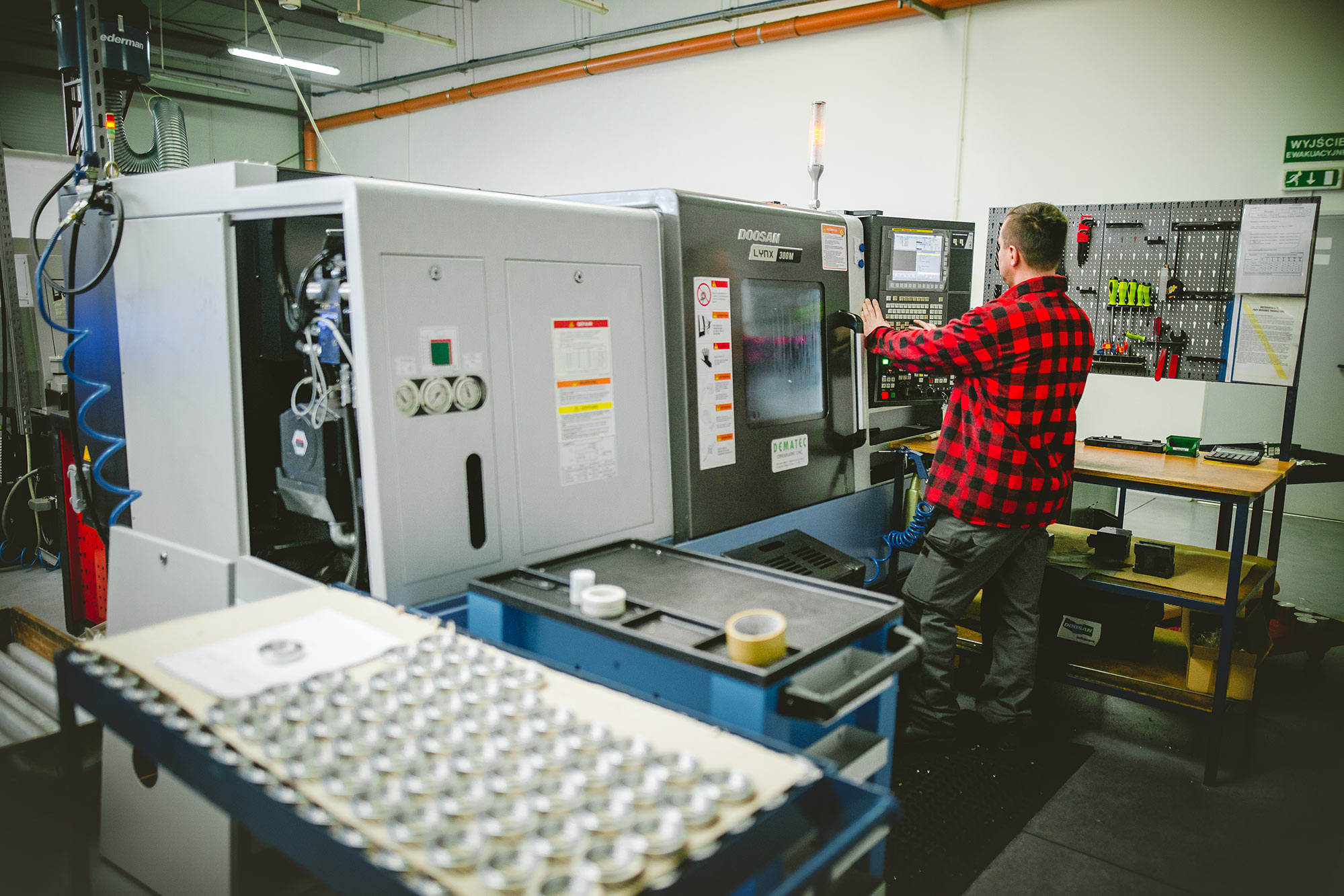 An employee at a DOOSAN Lynx 300M lathe in the shop floor - a tray of steel parts in the foreground
