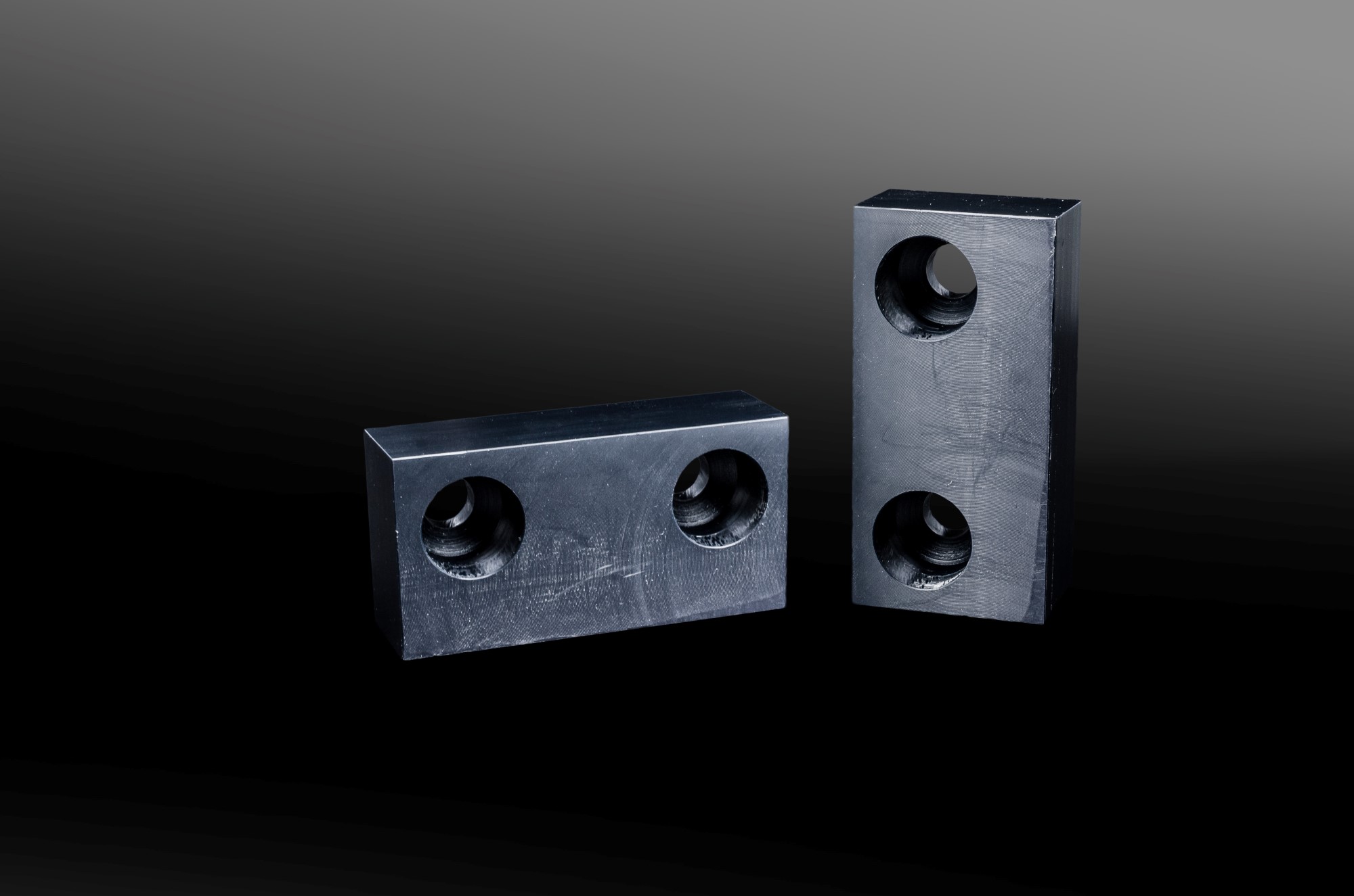 Two black, rectangular metal blocks with countersunk holes, set against a dark, gradient background.