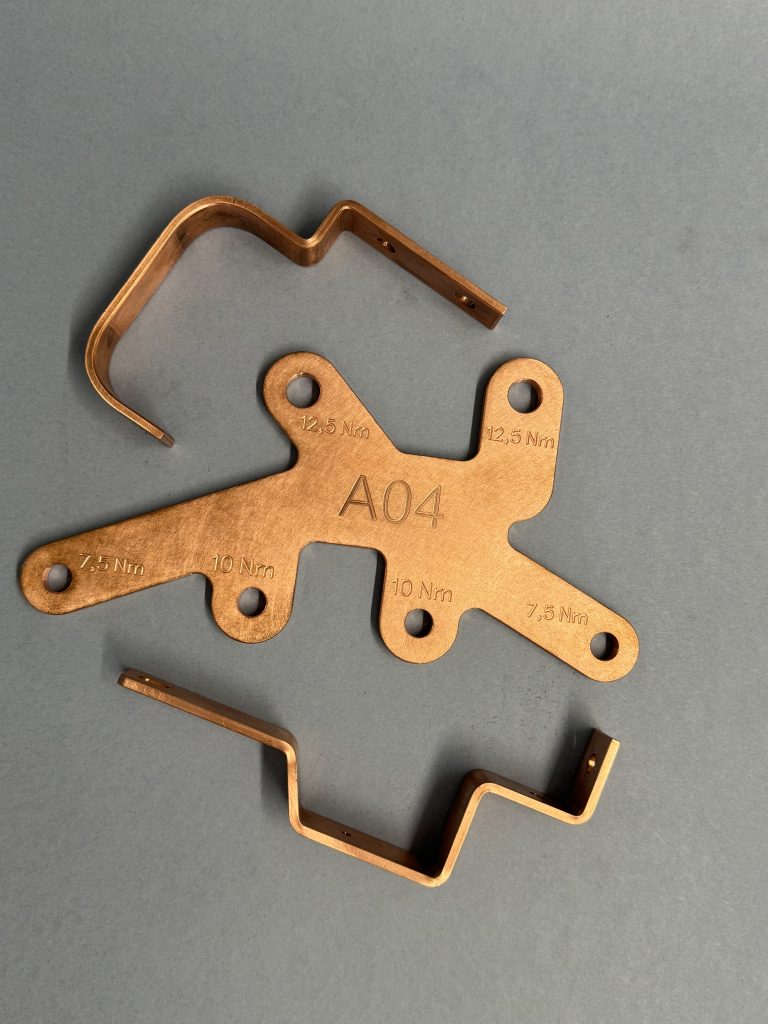 Three copper components on a grey background, including a flat plate marked A04 with Nm markings and two bent profiles.