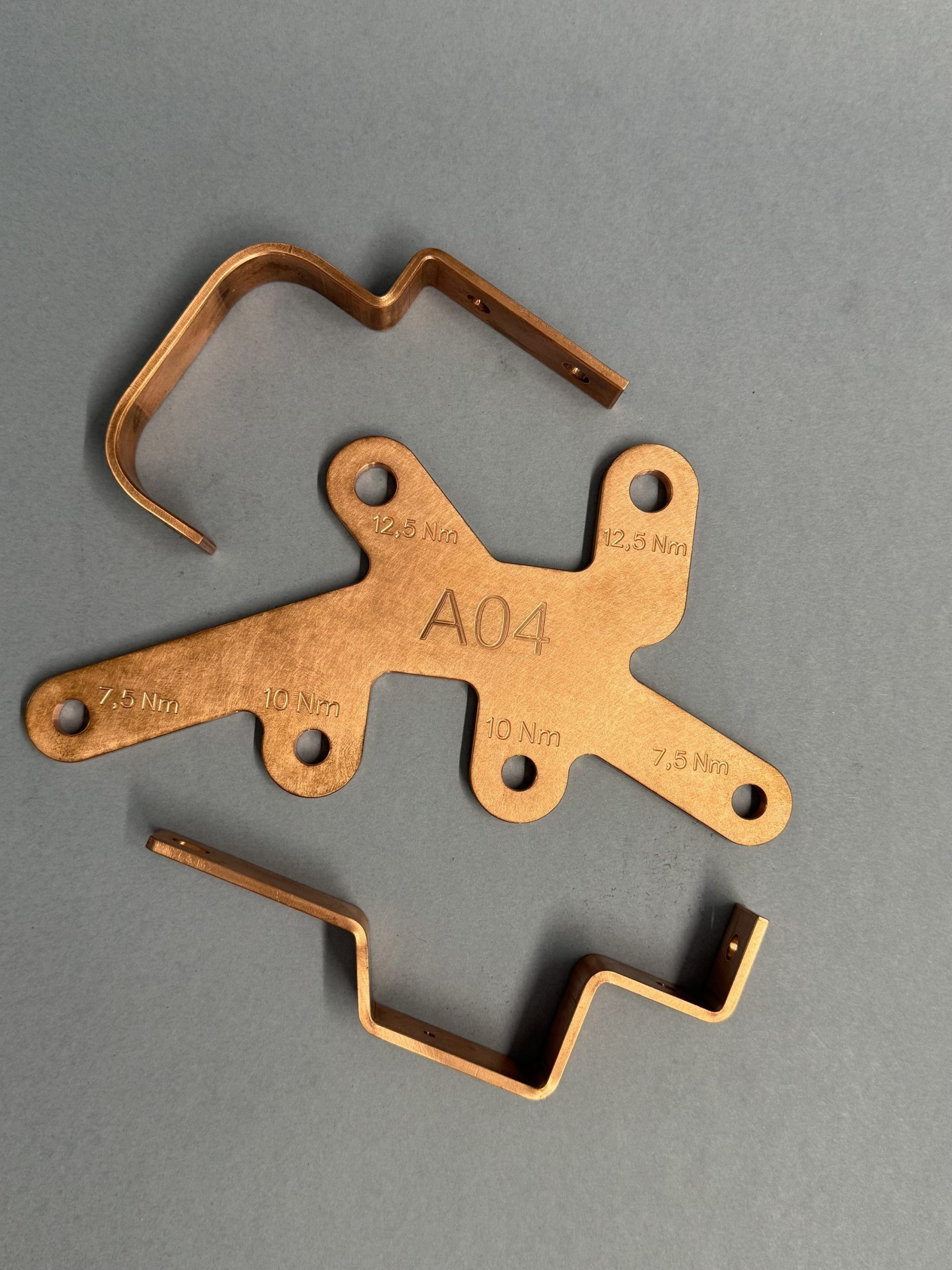 Three copper components on a grey background, including a flat plate marked A04 and Nm and two bent profiles.