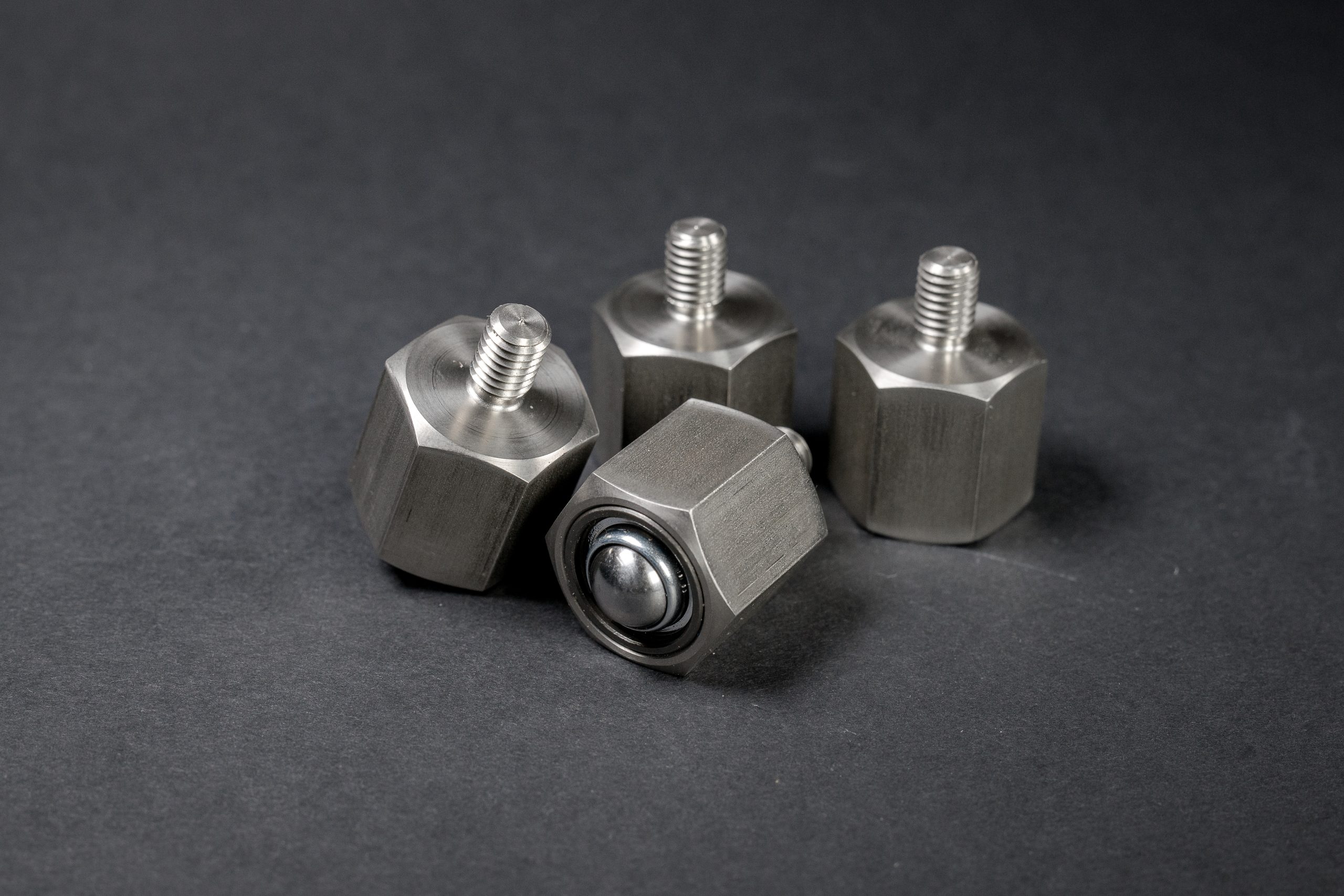 Four metal hexagonal components with a threaded stud and built-in ball bearing on a dark background.