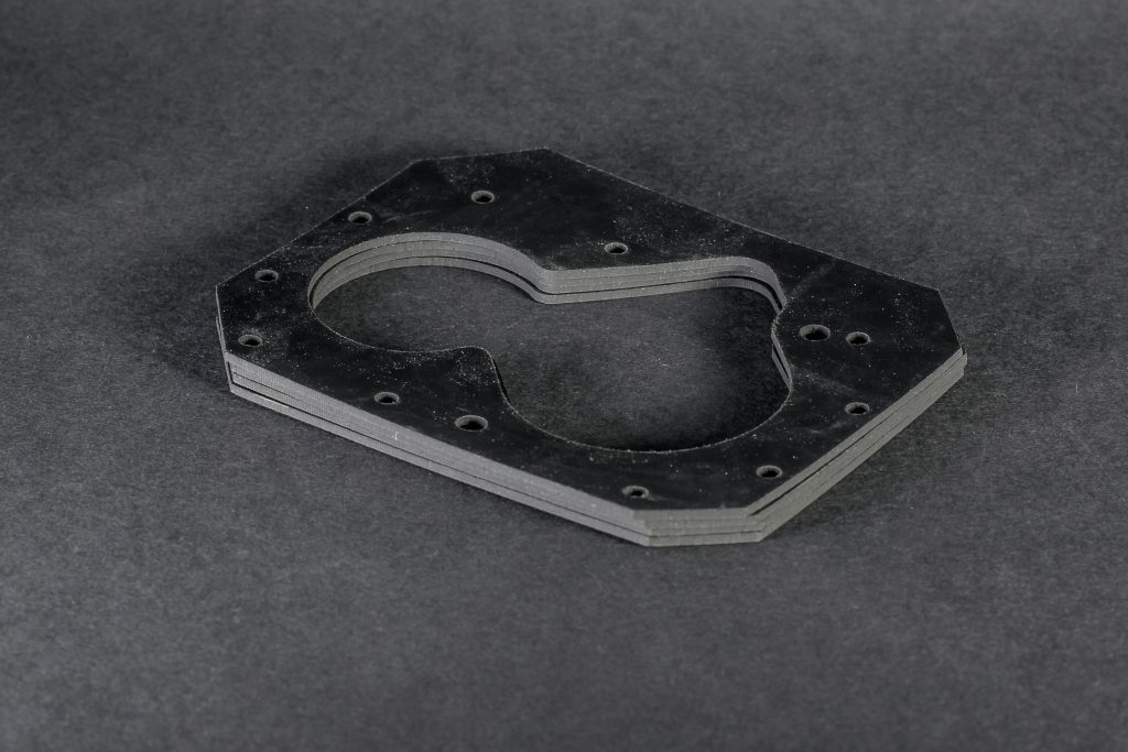 A stack of black flat technical gaskets with an irregular outline and bolt holes on a dark surface.