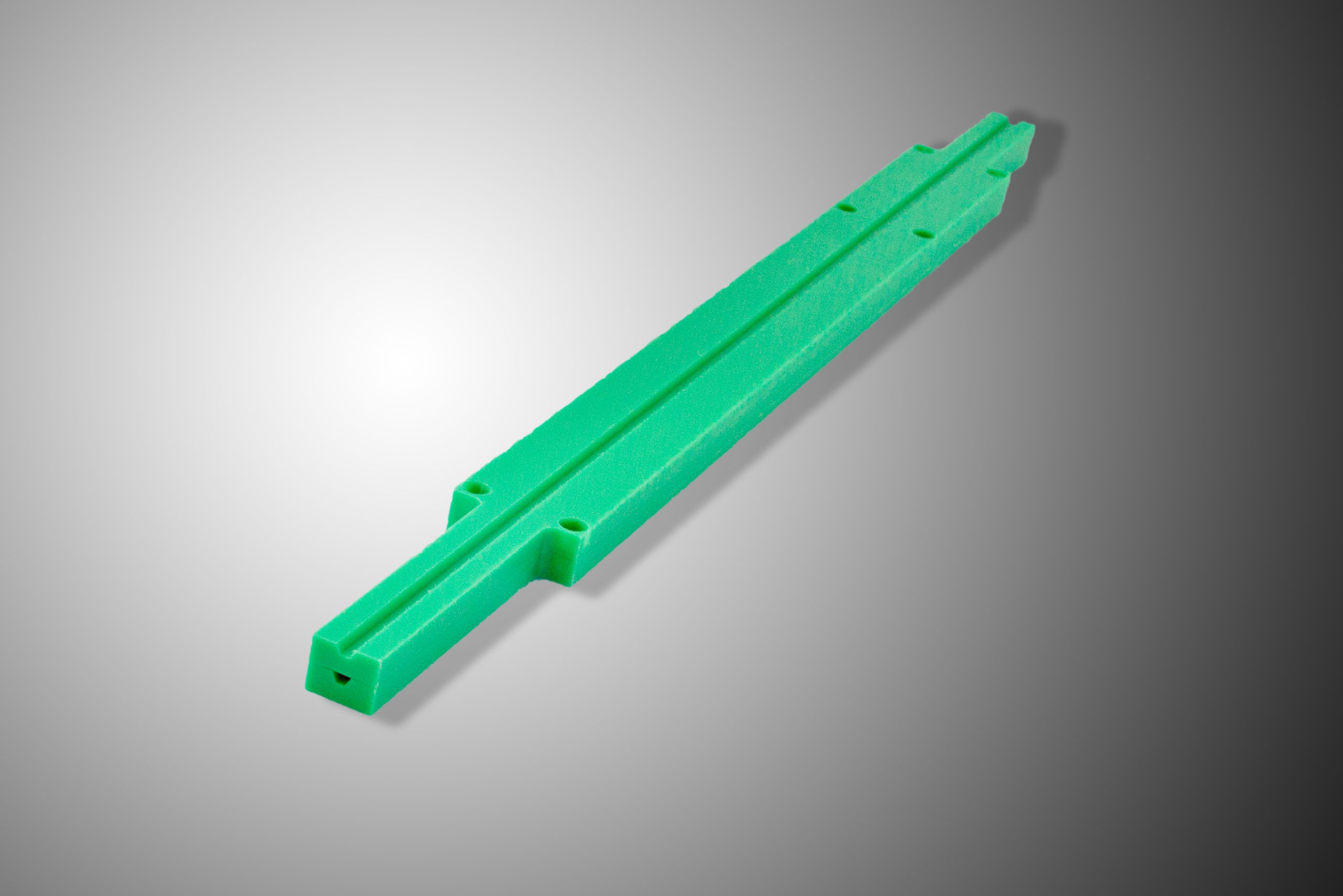 A green elongated part with a groove running along its whole length, holes and profiled tapers lying diagonally on a grey background.