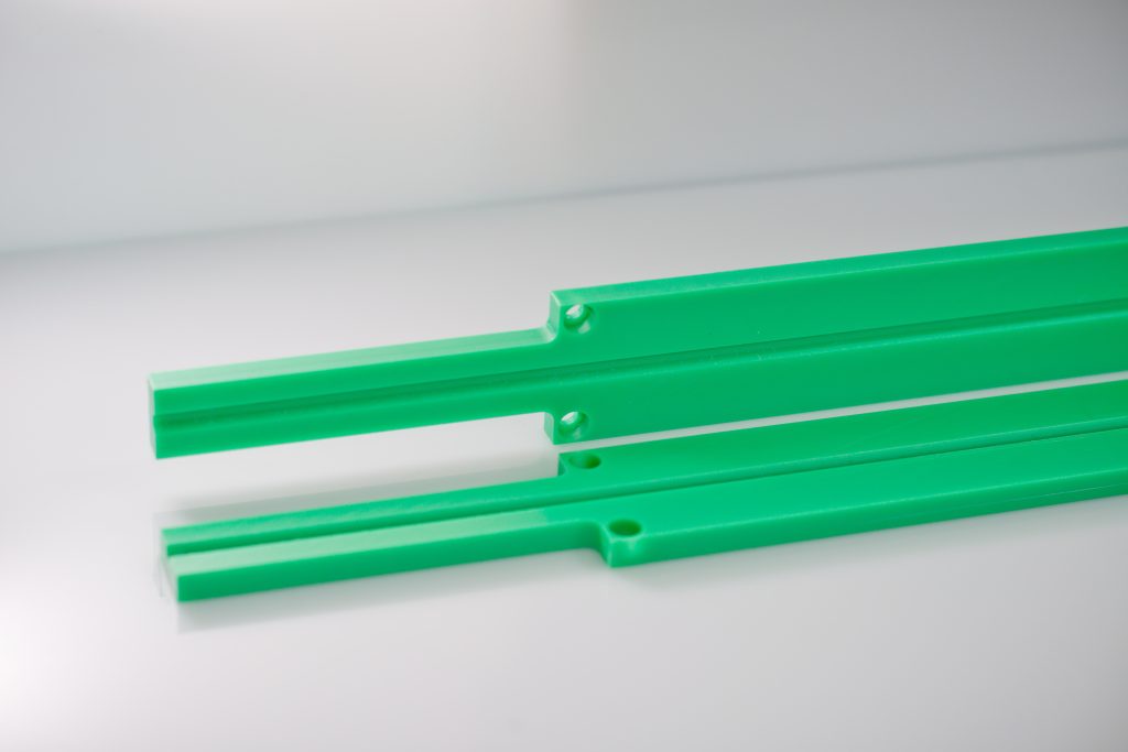 Two elongated green plastic guide rails with milled grooves and holes on a light surface.