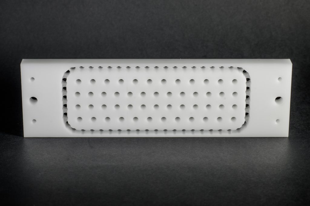 A white, rectangular part of smooth plastic with a central grid of regular holes and holes at the sides, on a background.