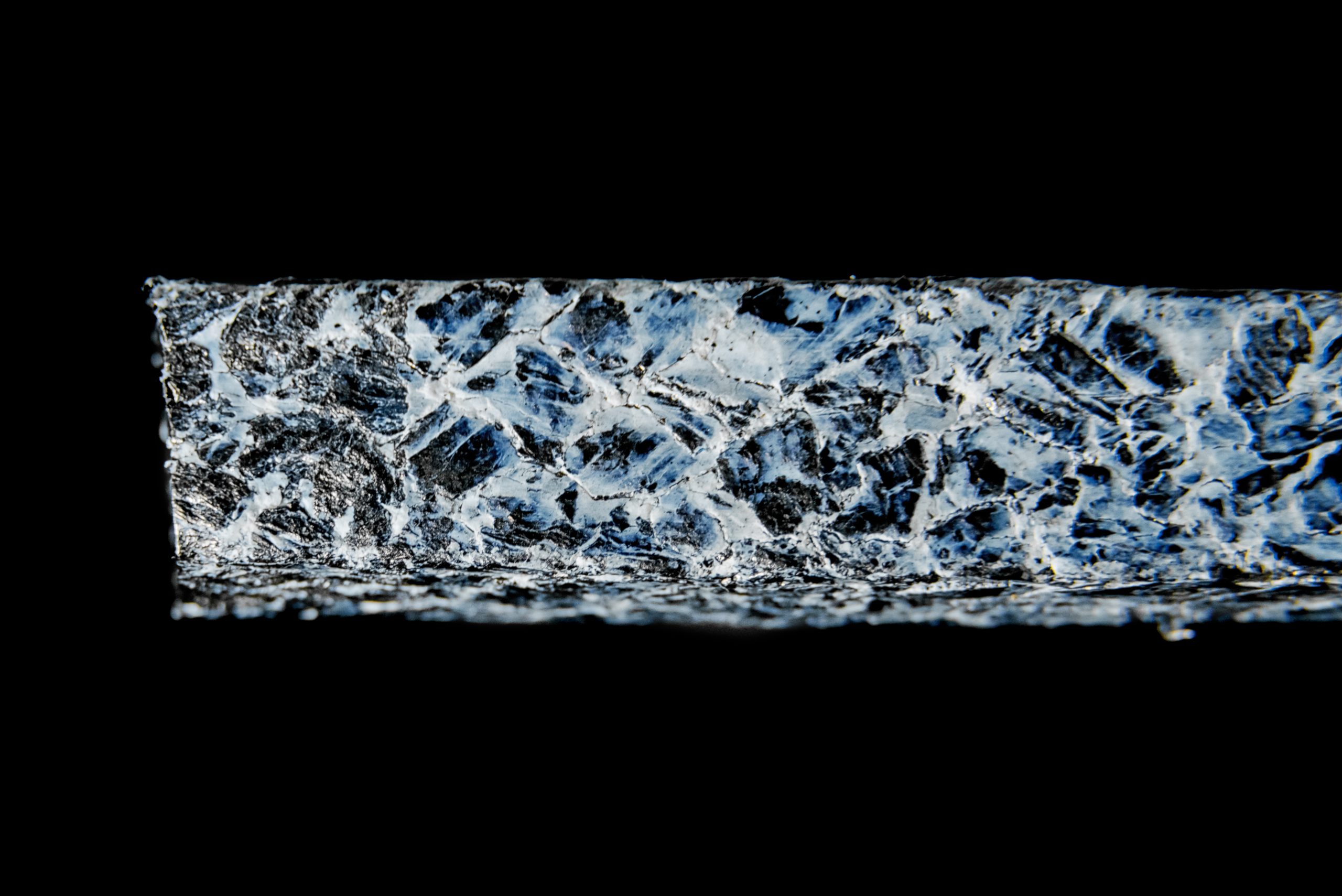 Strip of cracked sealing material with a white coating, shown at high magnification against a uniform black background.