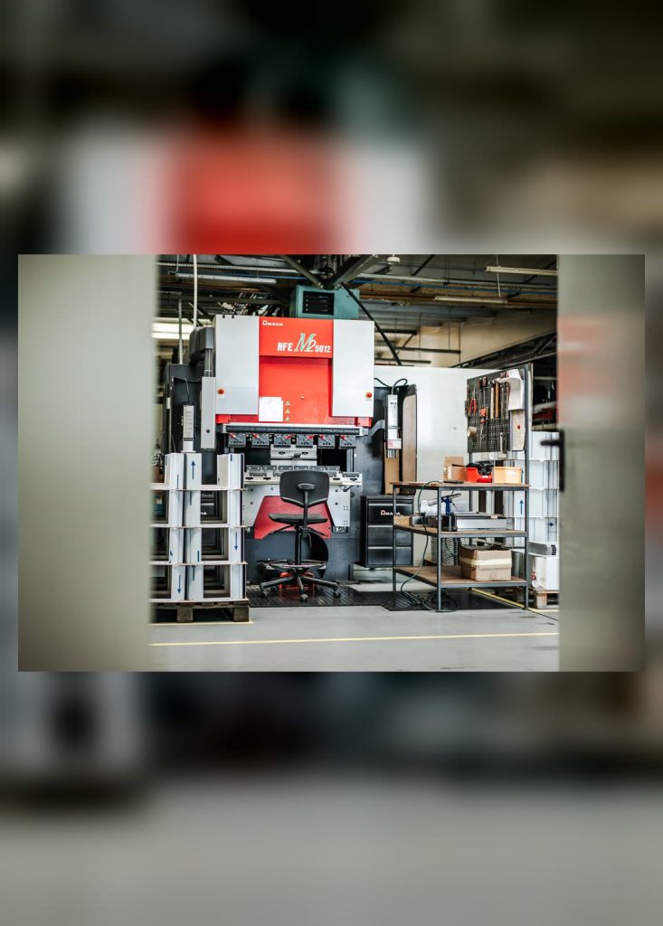 Red press brake in a production hall with a swivel chair, a tool trolley and pallets by the workstation.