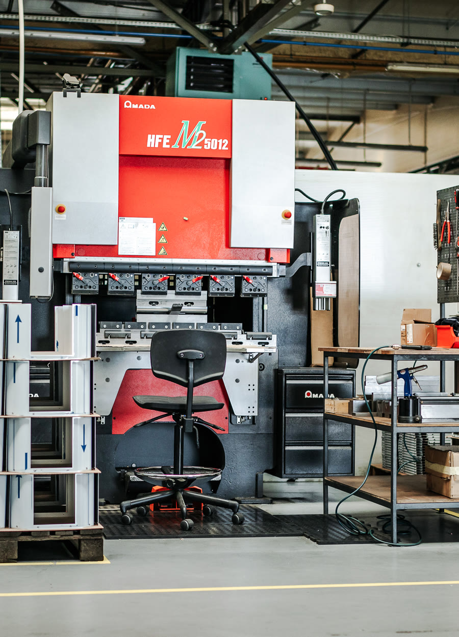 Red and grey press brake in an industrial hall with a black swivel chair and a pallet of bent profiles.