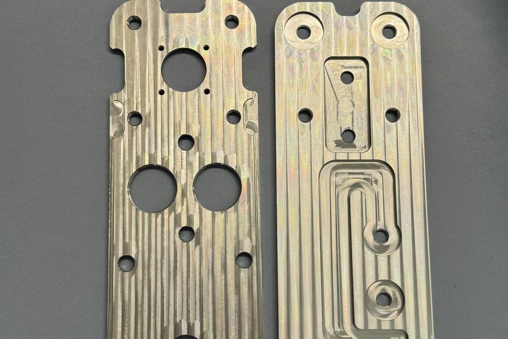 Two milled metal components with numerous holes and grooved channels, lying side by side on a grey background.