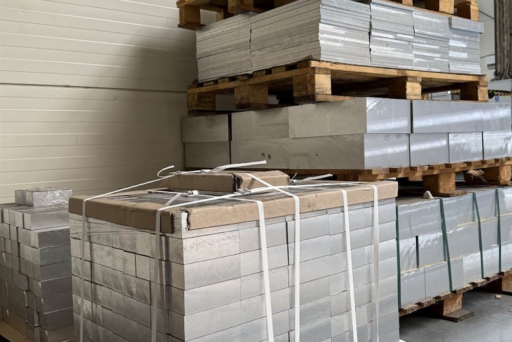 Stacks of metal blocks and aluminium plates strapped with plastic bands, arranged on wooden pallets in a warehouse.