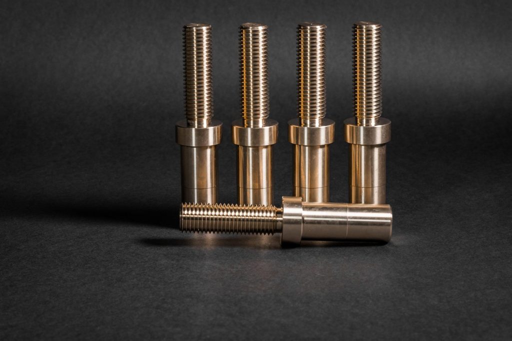 Five threaded brass pins with a flange, four standing upright and one lying across them against a dark background.