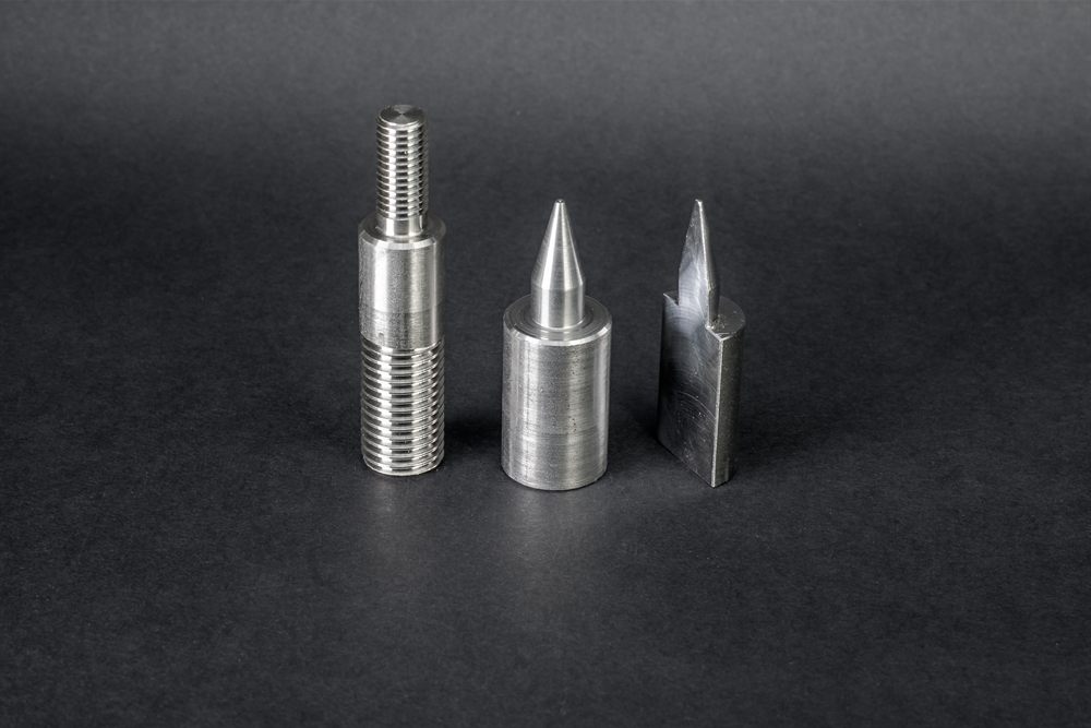 Three machined metal parts of various shapes, including one with a thread and a conical tip, standing against a black background.