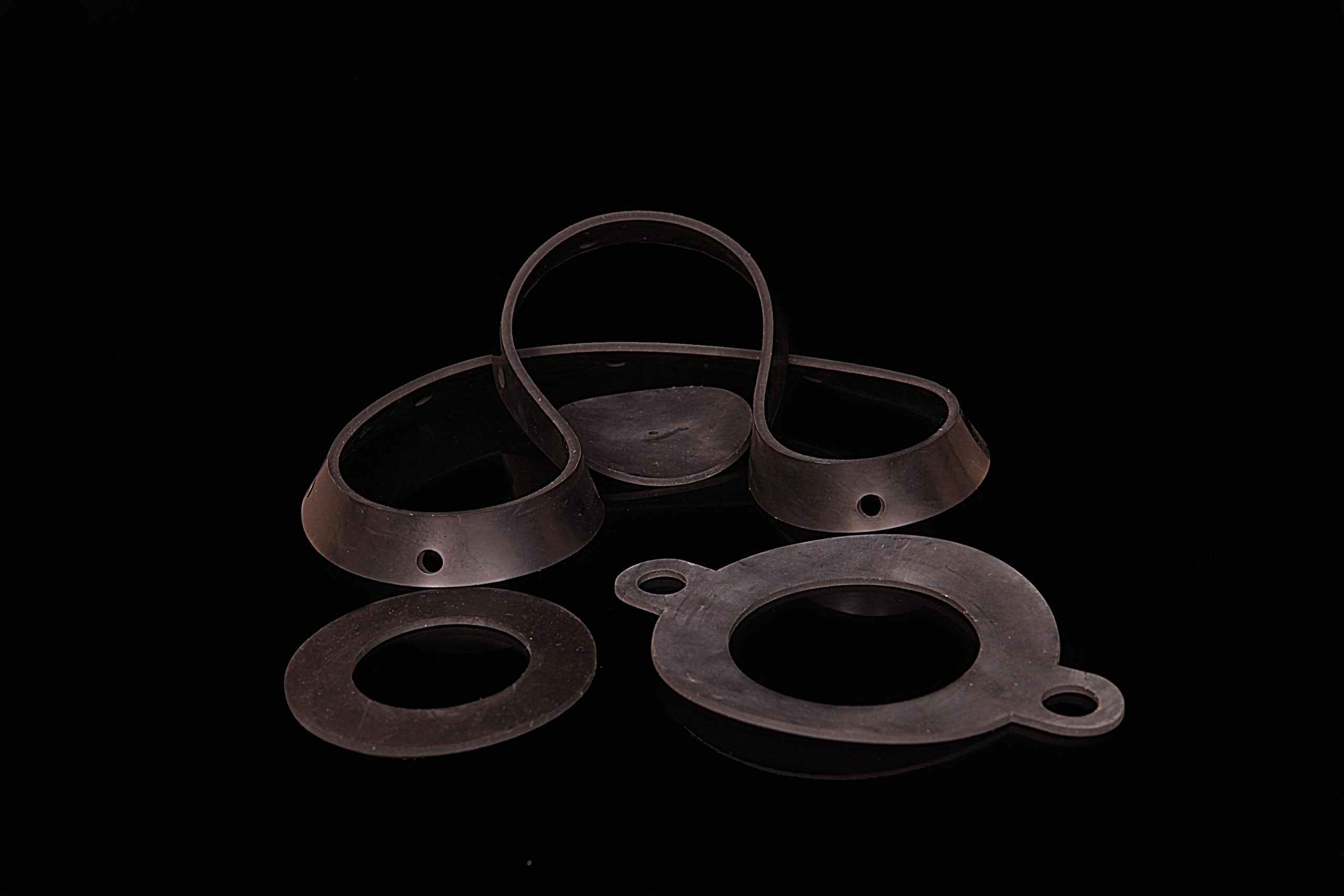 Black flexible gaskets of various shapes and with various holes lying on a glossy worktop against a dark background.