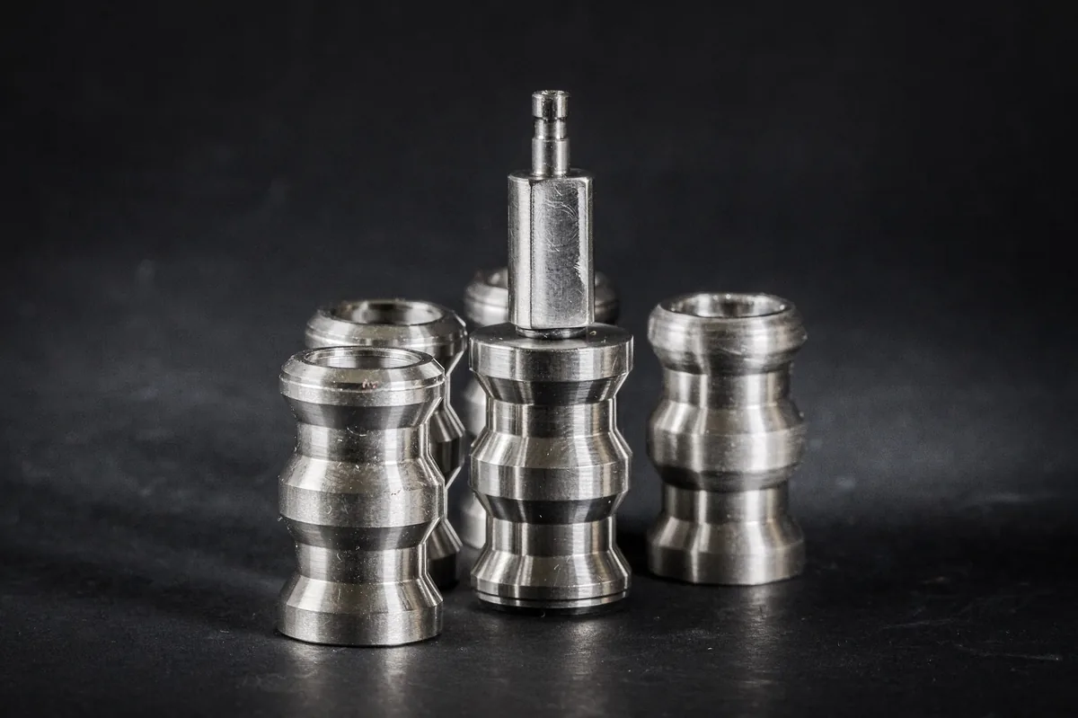 Five silver cylindrical components with profiled grooves, standing on a black, matte surface.