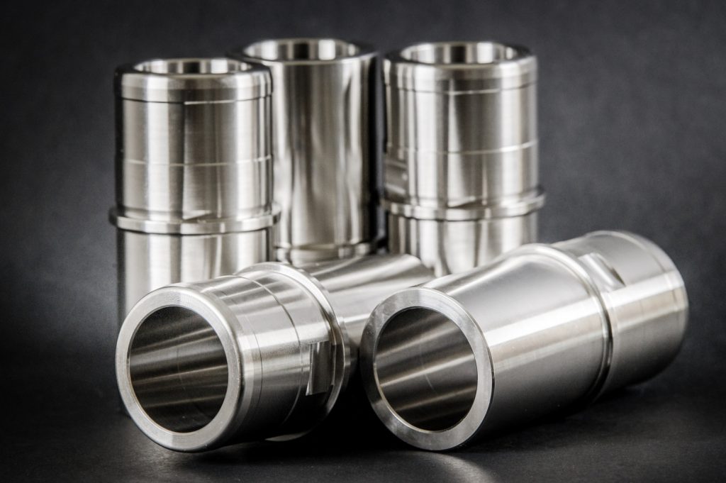 Five precisely turned cylindrical metal sleeves with grooves, three standing and two lying against a dark background.