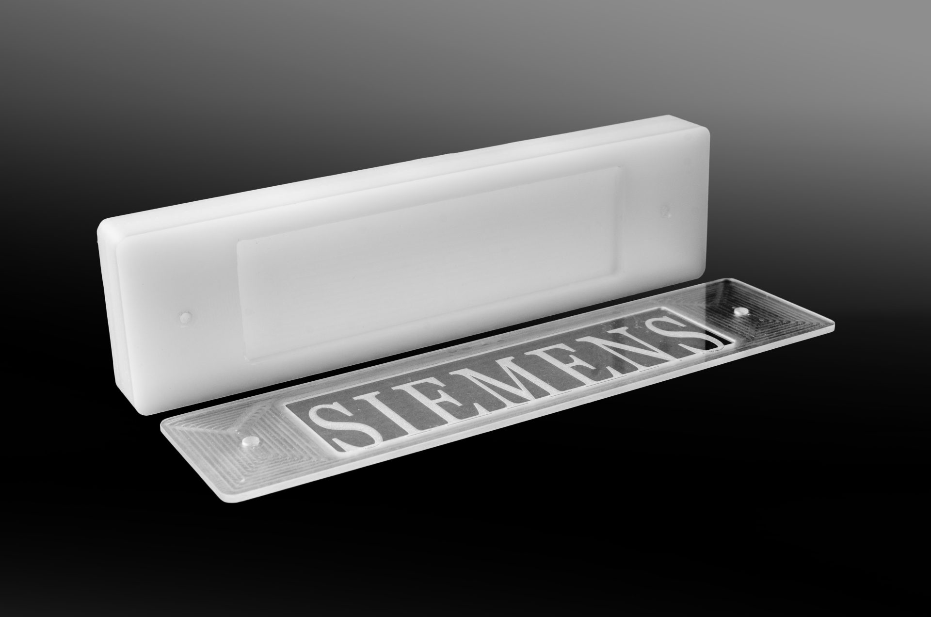 White plastic block with a transparent plate bearing a milled inscription lying in front of it on a dark background.