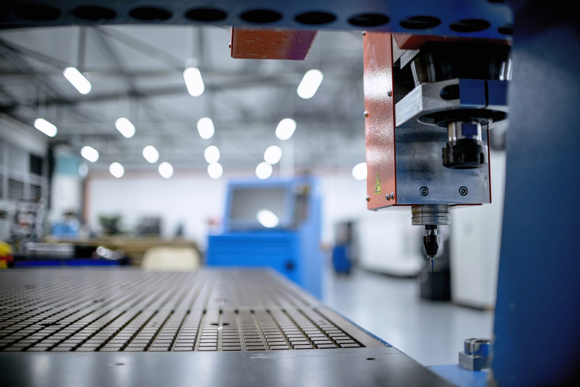 A red milling machine head with a metal workpiece hangs above the table, with rows of ceiling lamps in the background.