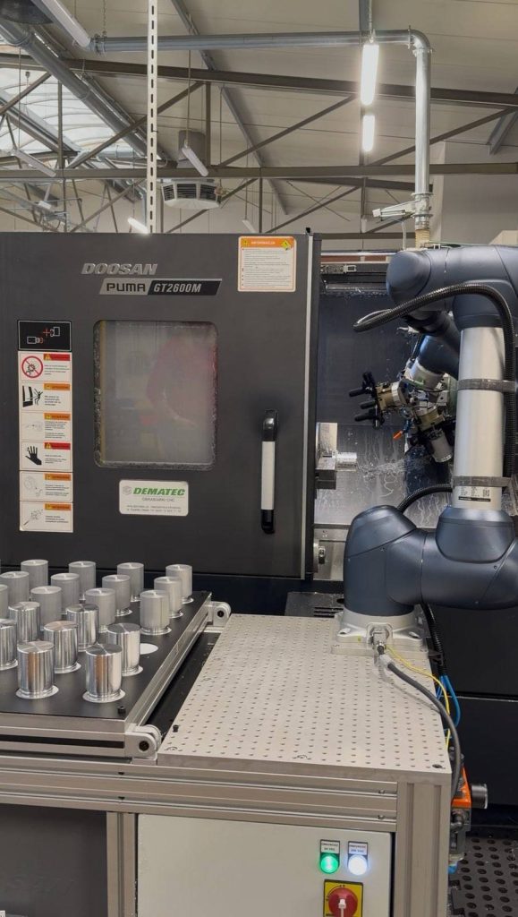 Tokarka CNC Doosan PUMA GT2600M i ramię robota obok tacy z metalowymi, cylindrycznymi detalami w hali.
