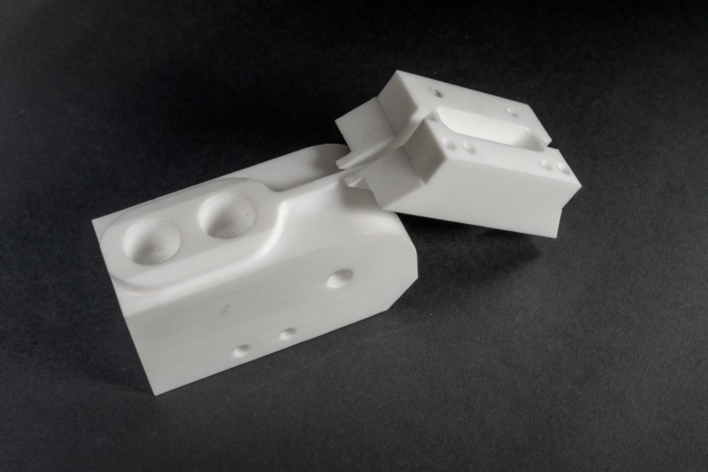 Two white milled plastic parts with special seats and holes on a dark background.