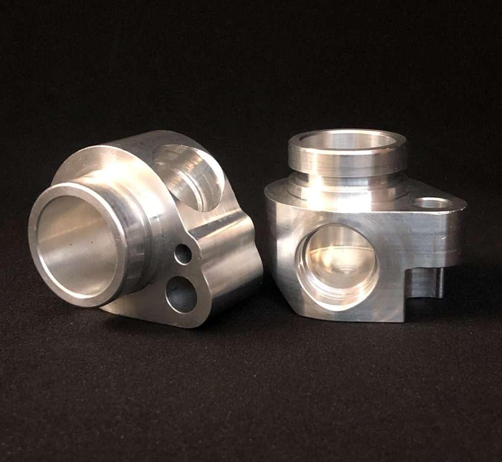 Two precisely machined metal bodies of complex shape with cylindrical chambers standing against a black background.
