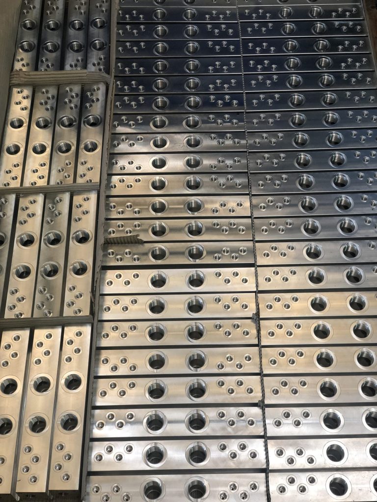 Rows of rectangular metal blocks with numerous holes and threads stacked on a pallet and secured with straps.