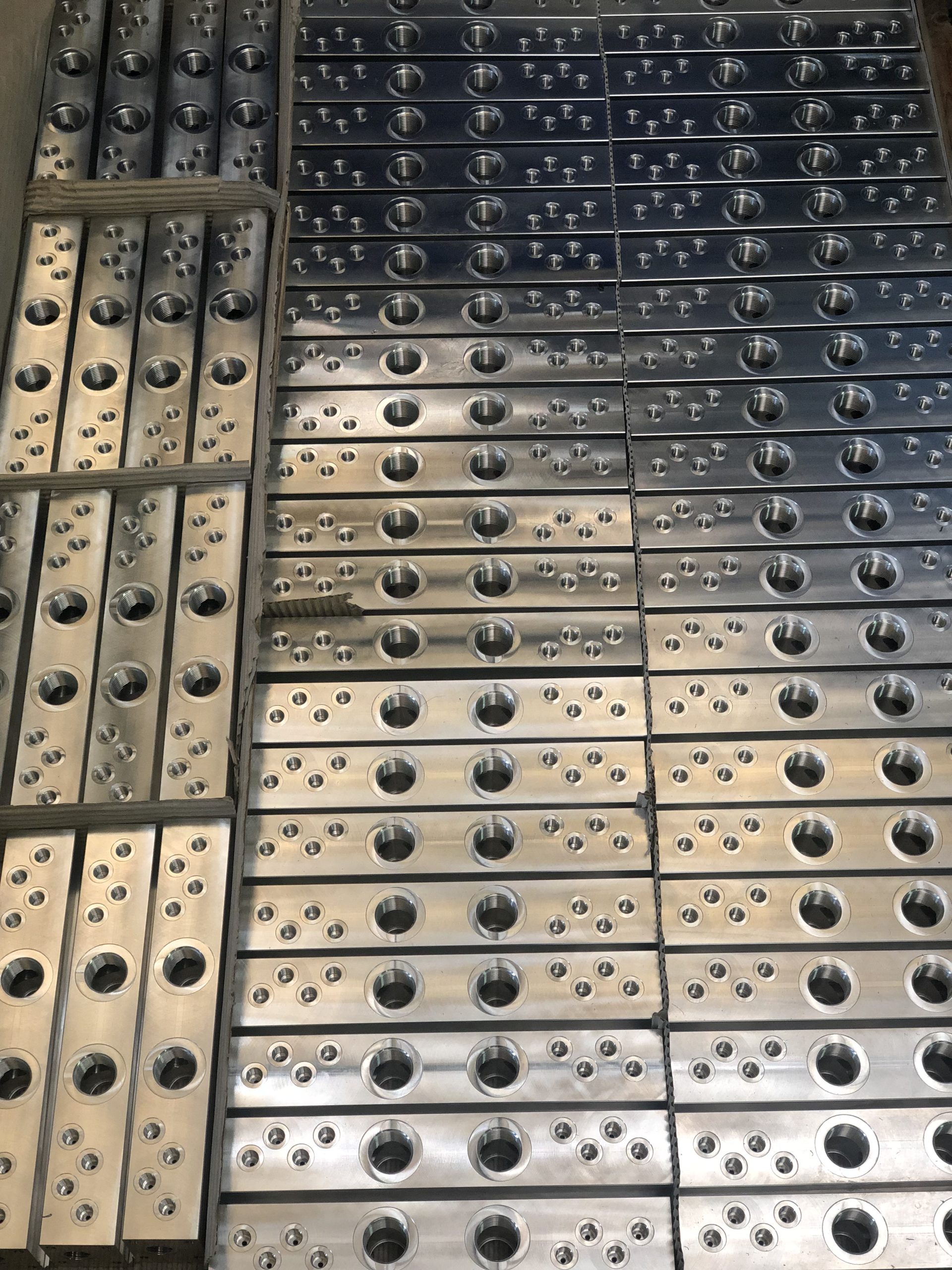 Rows of rectangular metal blocks with numerous holes and threads arranged on a pallet and secured with straps.