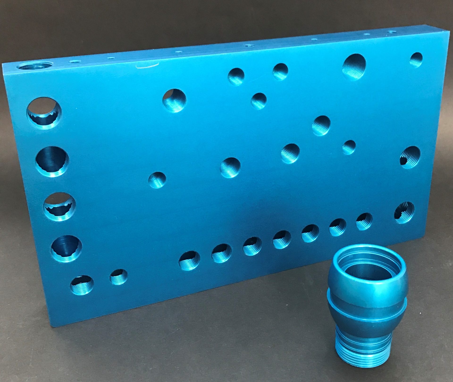 Blue rectangular plate with numerous threaded holes and a small cylindrical detail standing against a dark background.