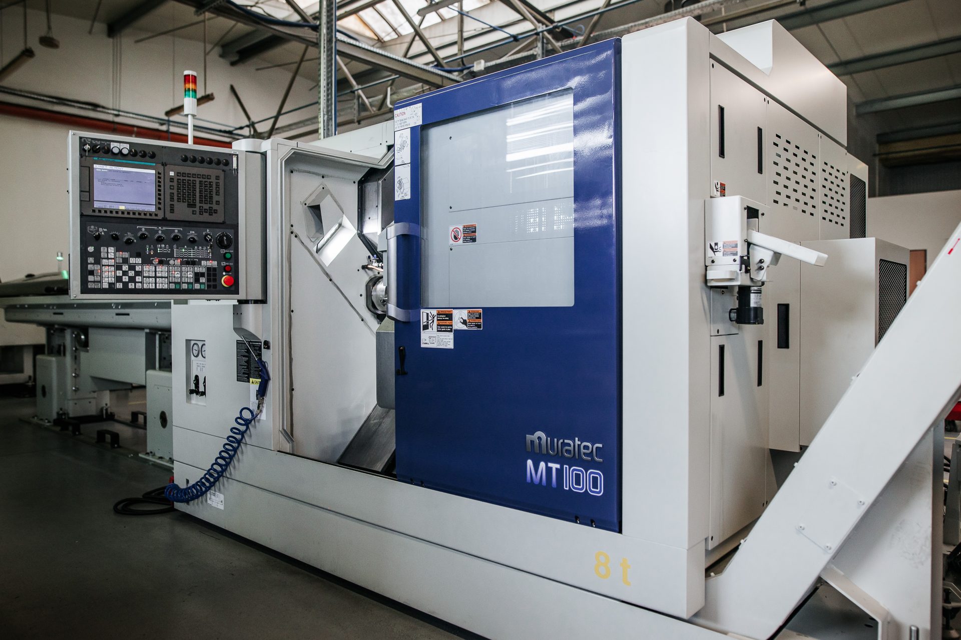 A modern CNC machine tool with a white and blue panel stands on a grey floor in a bright production hall.