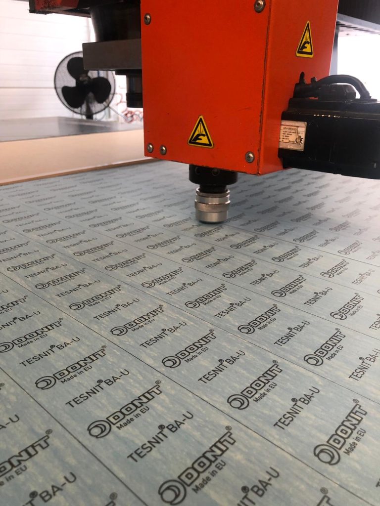 An orange machine cutting head with yellow warning signs above a blue sheet of material.