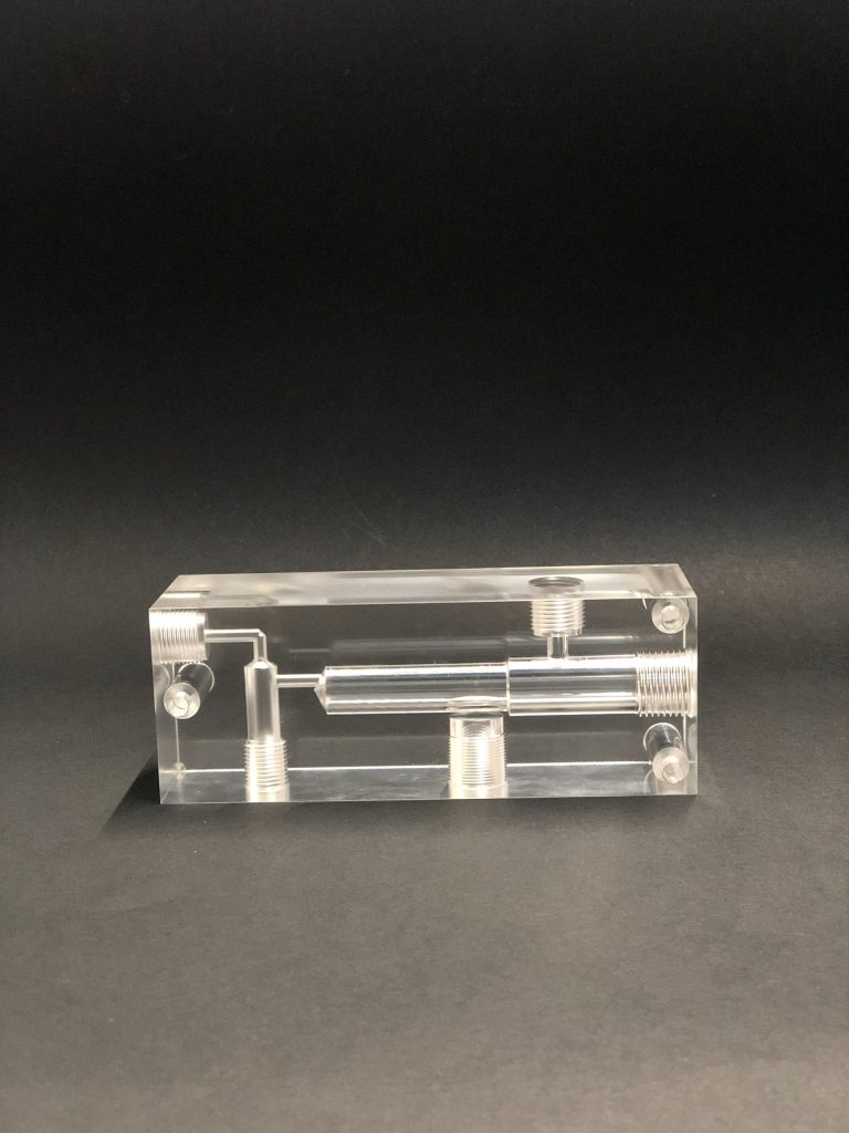 Transparent rectangular block with internal channels, threaded holes and four screws in the corners, on a dark table.