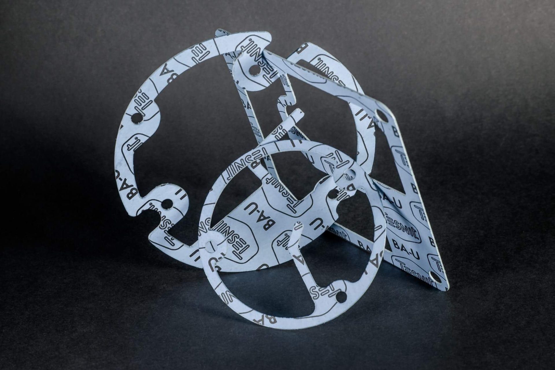 A sculpture made of light grey cut-outs in various shapes, with markings and holes, interwoven into a solid form against a black background.