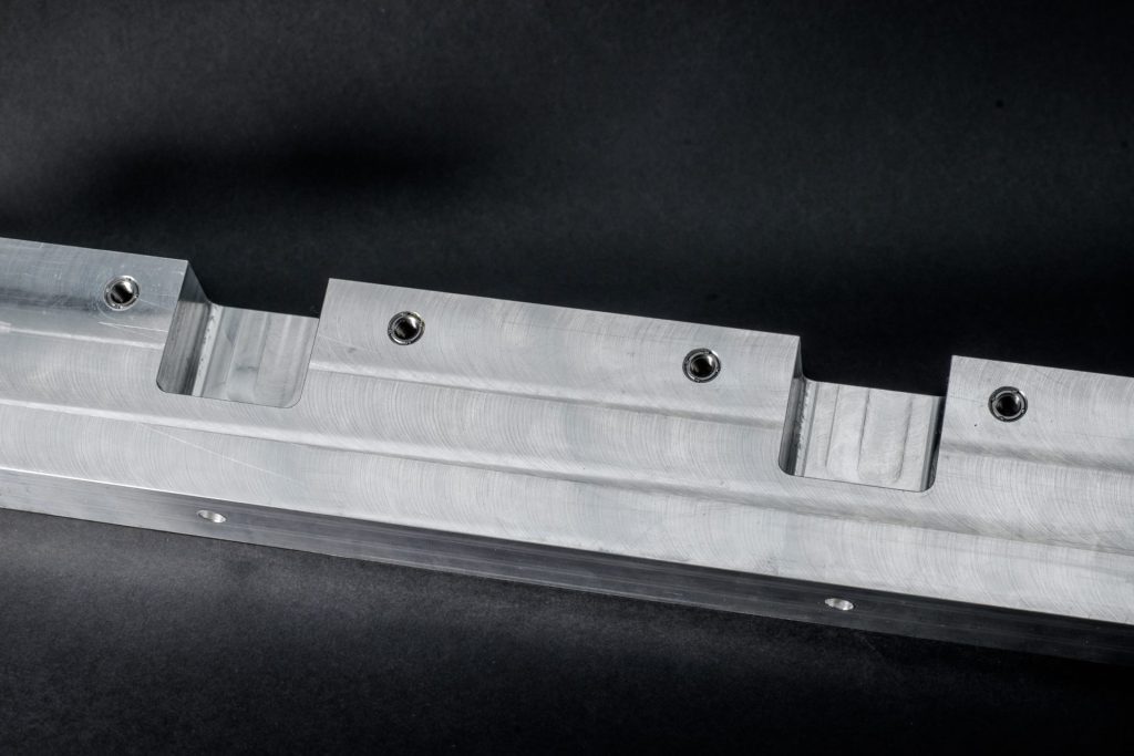 Silver-grey metal profile with milled recesses and four threaded bushings on a dark background.