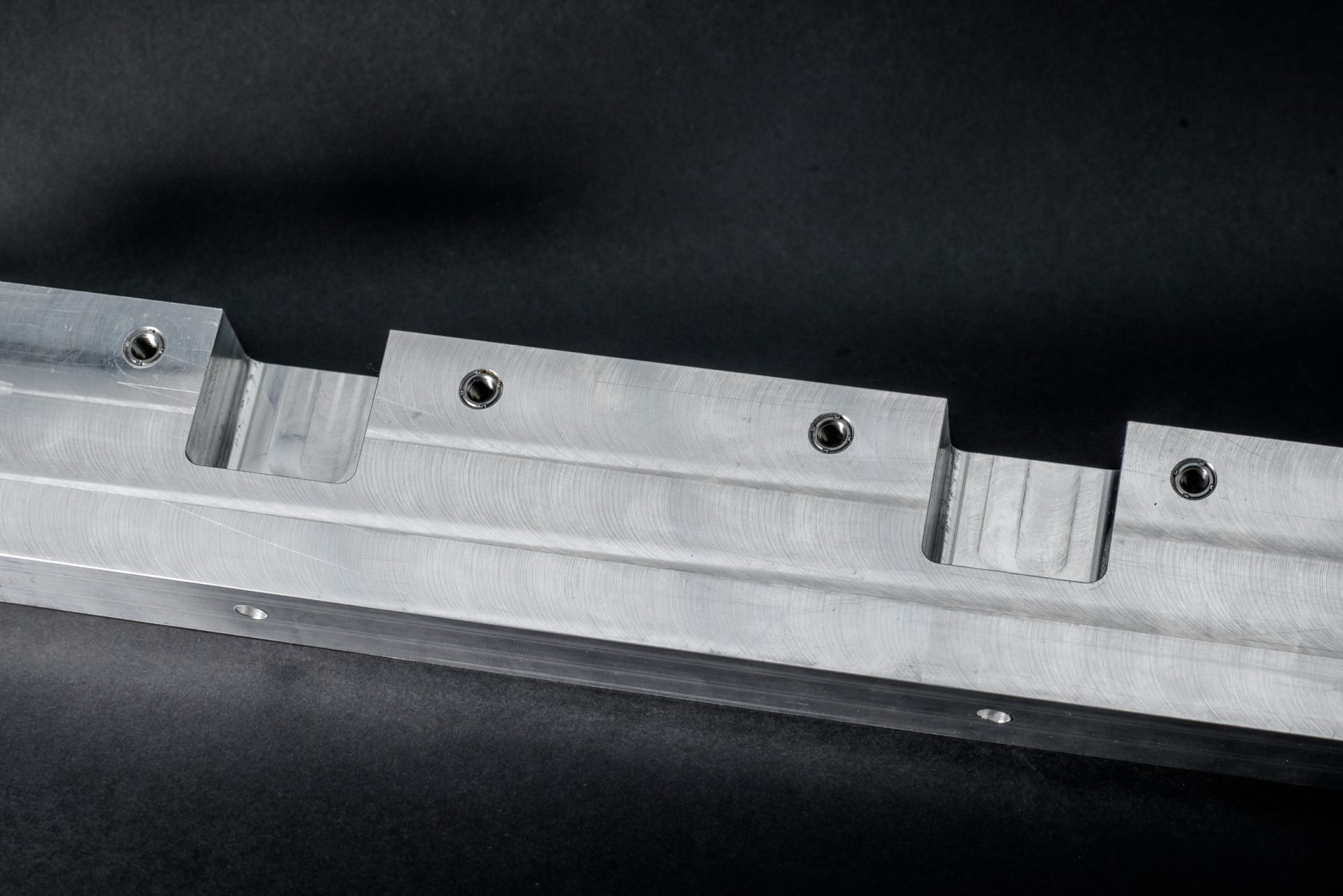 A silver-grey metal profile with milled recesses and four threaded inserts against a dark background.