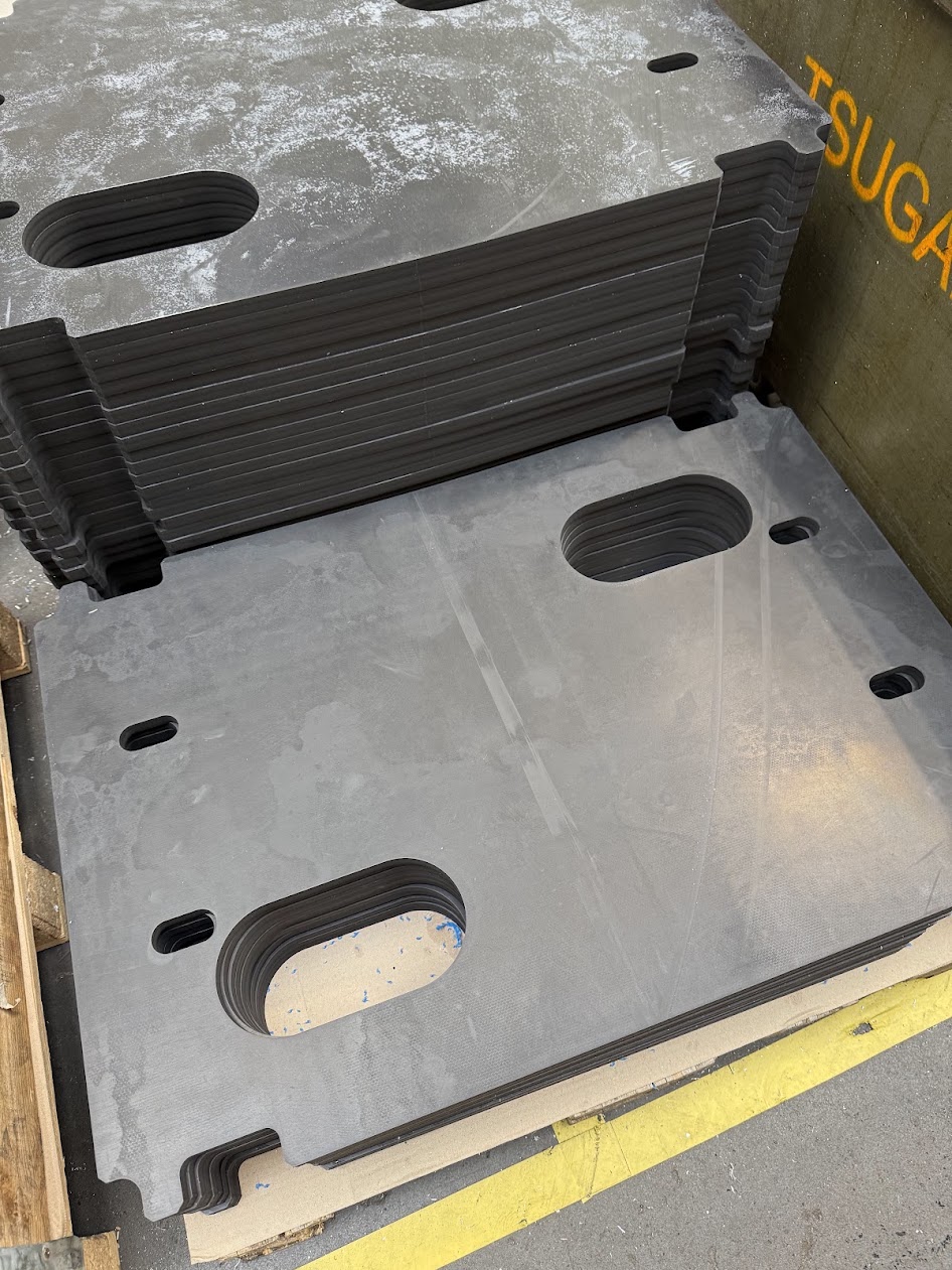 Stacks of dark grey technical plates with cut-out elongated holes arranged on a wooden pallet in a workshop hall.
