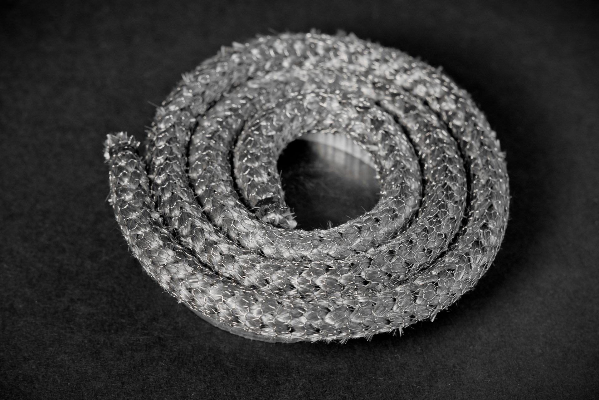 A silver-grey braided cord coiled in a spiral, with a fibrous rough texture against a dark matt background.