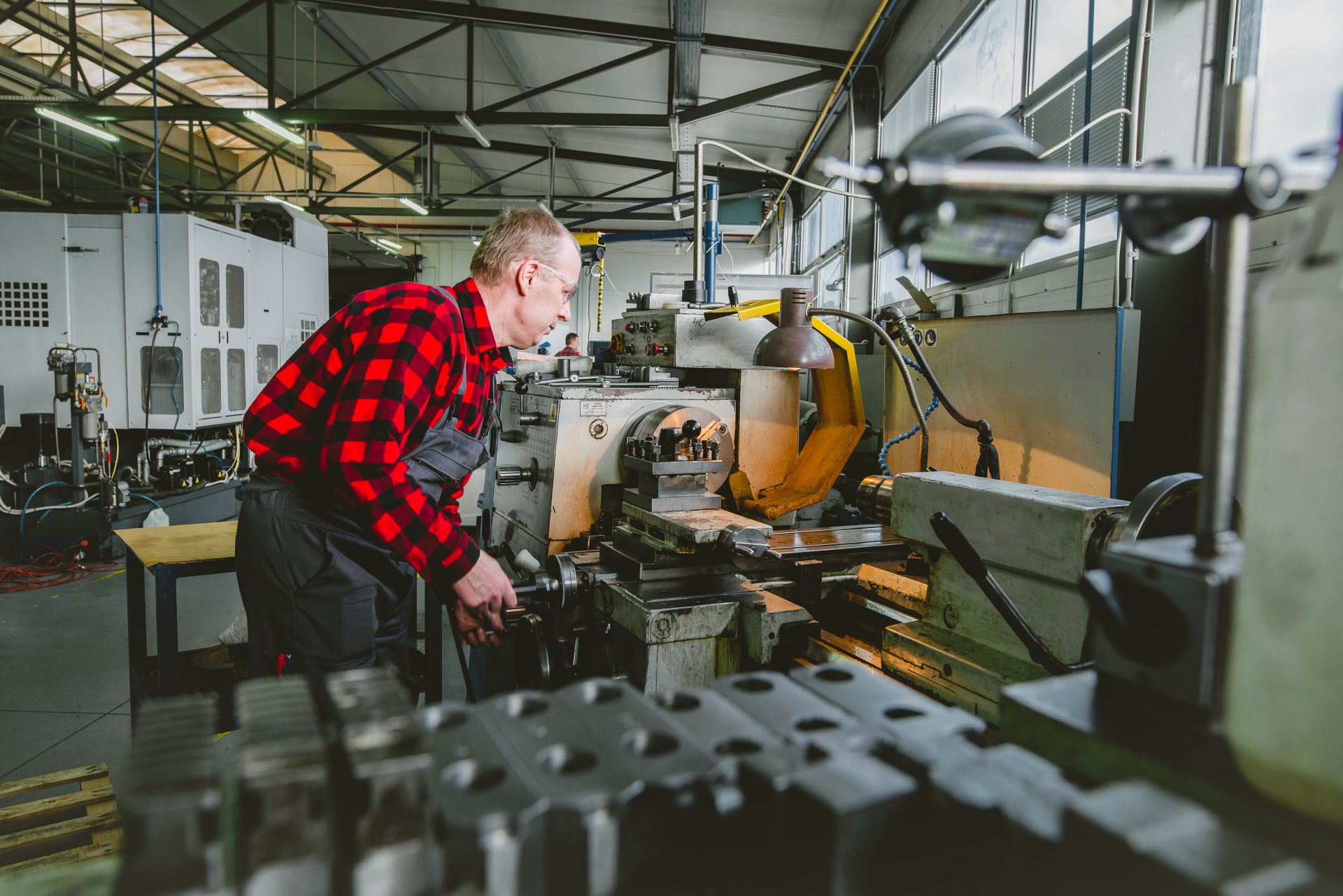 A worker in a red checked shirt operating a lathe with handwheels and levers in a brightly lit production hall.