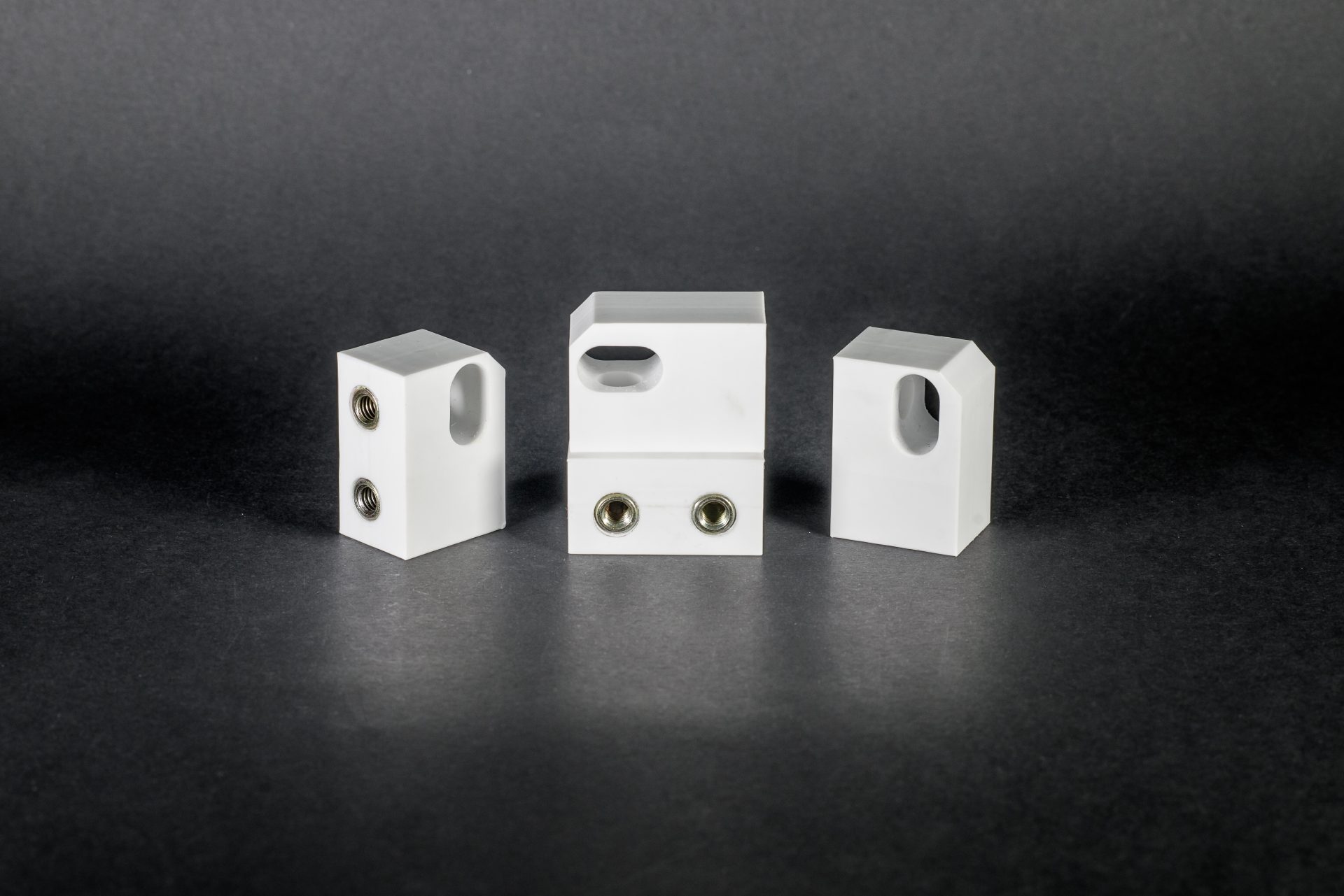 Three white plastic components with threaded inserts and holes arranged in a row against a dark background.