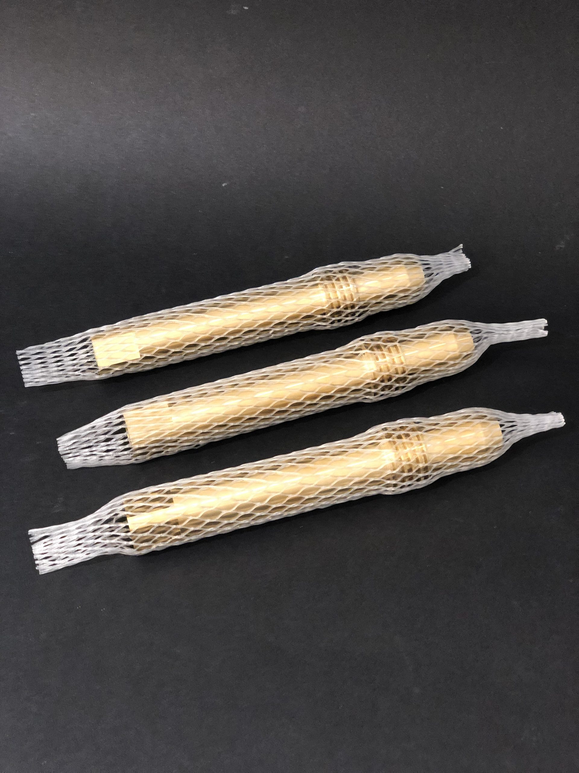 Three elongated gold-coloured items with a metallic sheen, lying diagonally on a black background, each in a separate white mesh sleeve.