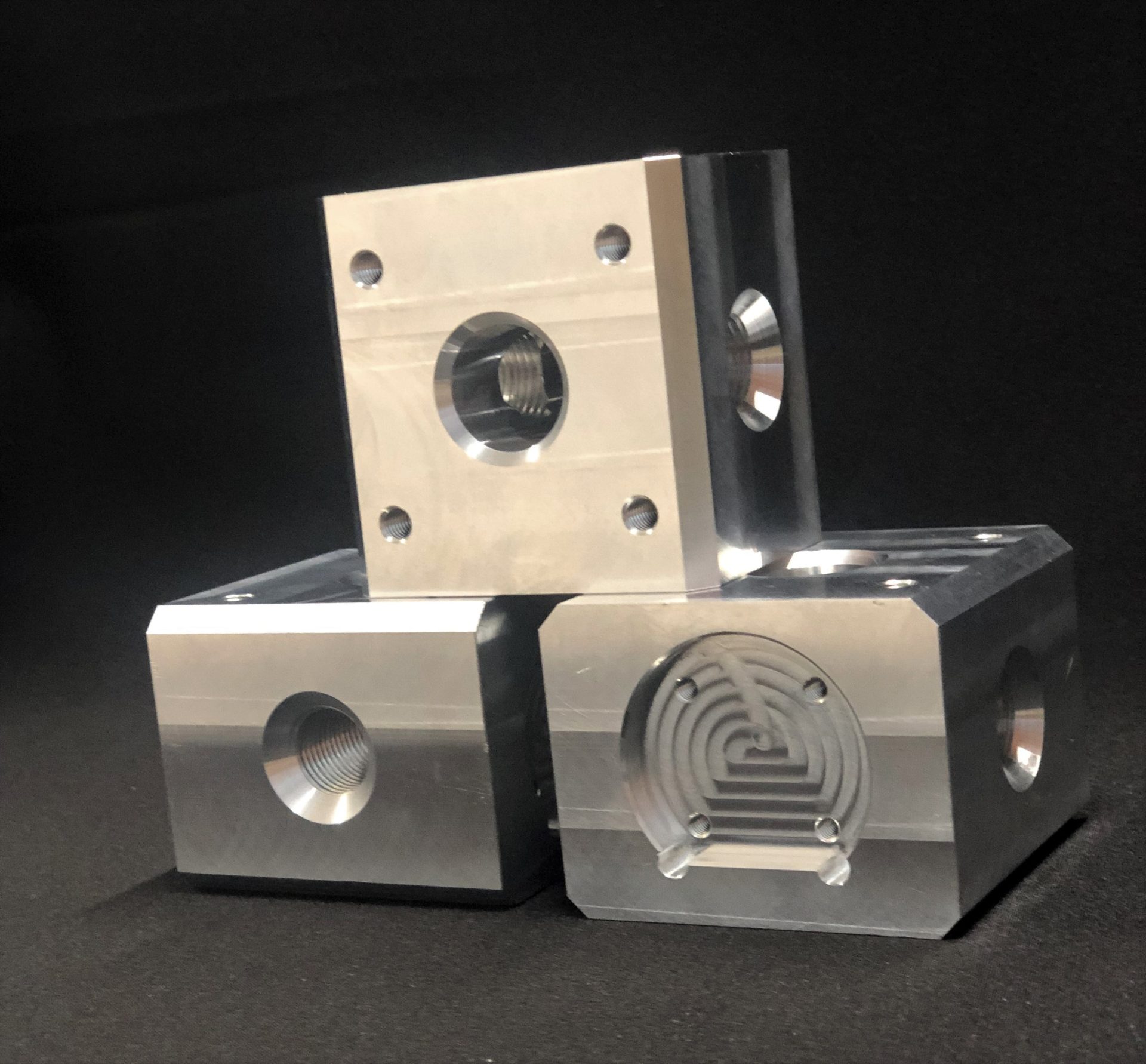 Three silver metal cubic blocks stand against a dark background; they have threaded holes and a milled pattern.