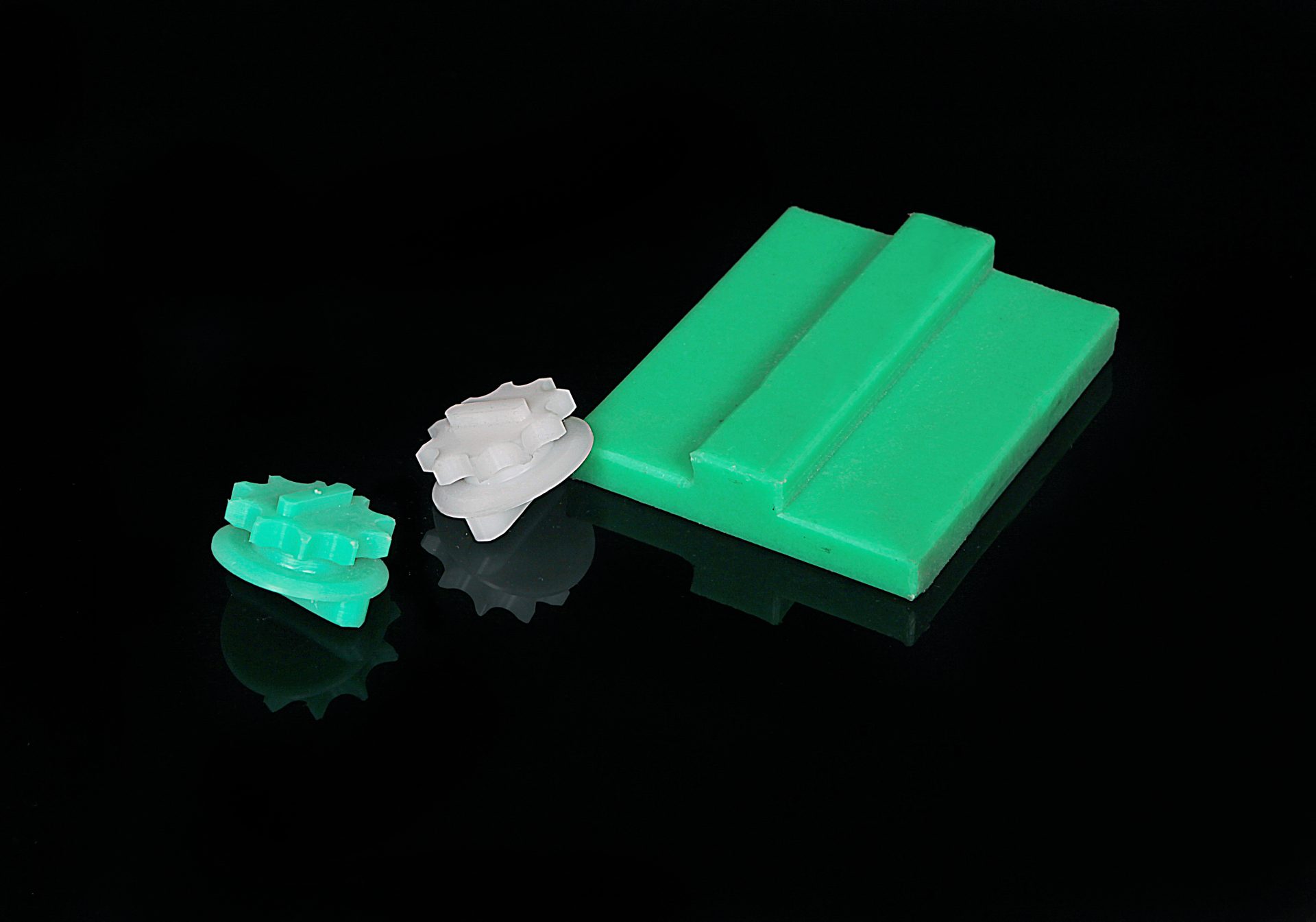 Green block with an elongated projection and two small parts with toothed gears, one green and one white, on a mirrored background.